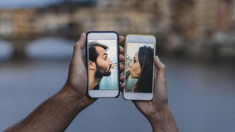 Dois telemóveis ao lado um do outro, cada um com uma foto de uma pessoa (mulher e homem) voltados um para o outro, a mandar um beijo